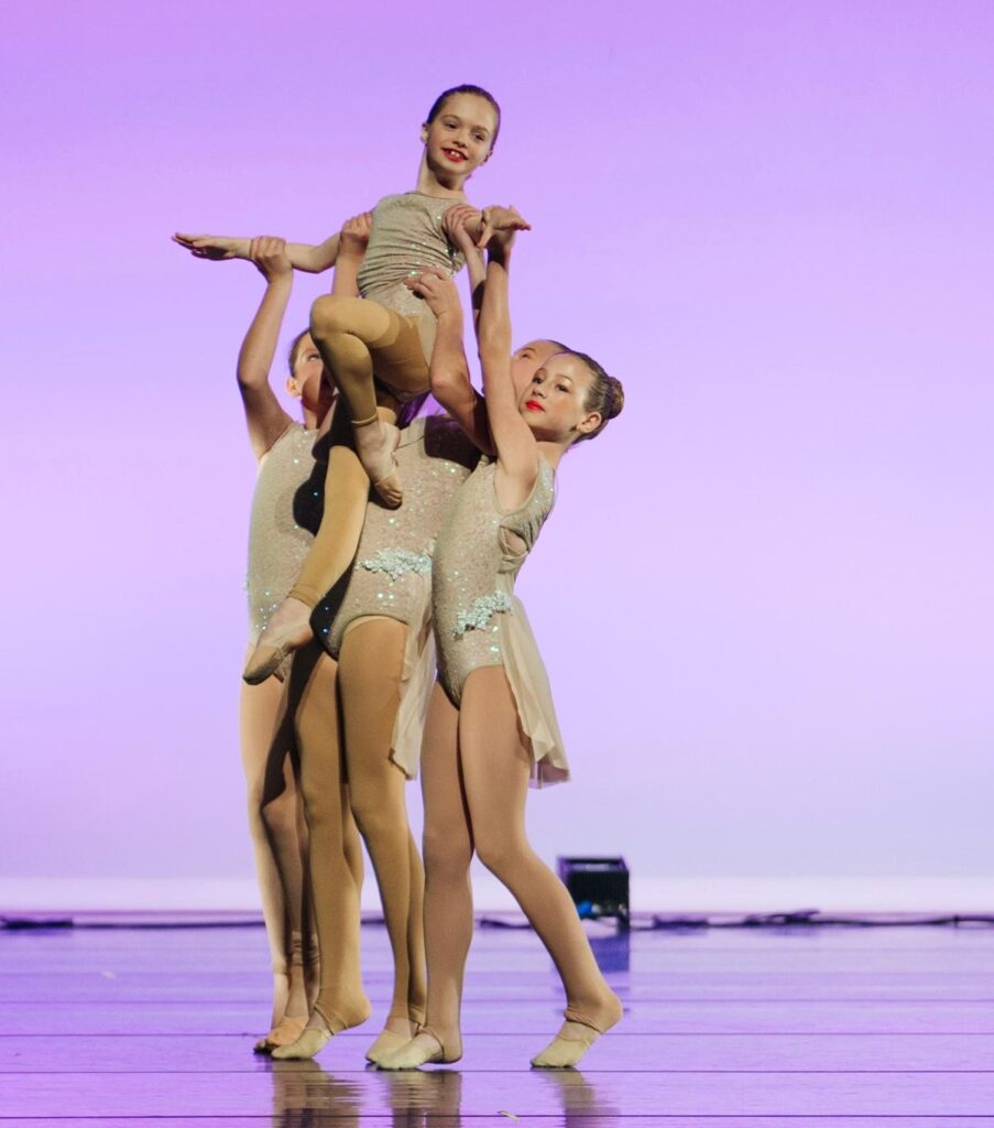 A group of young girls performing a ballet routine with grace and precision, taught by a RAD Certified Instructor. The girls are dressed in ballet attire, demonstrating techniques they learned through expert guidance in a bright, professional studio.